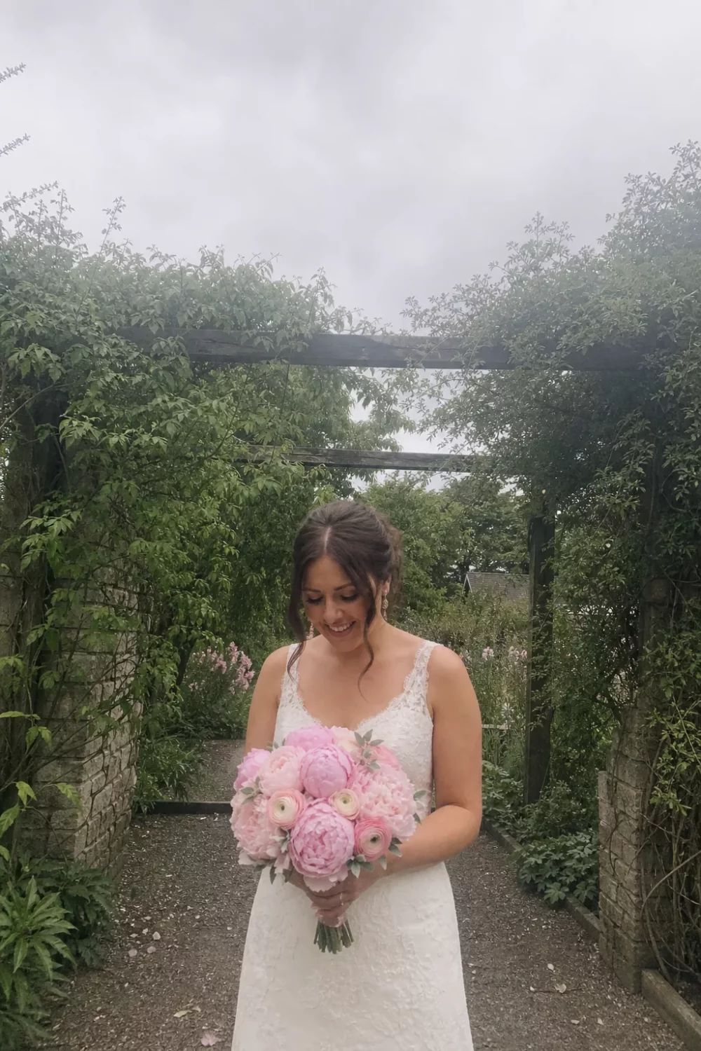 Bride holding a soft pink peony and ranunculus wedding bouquet in a garden arbor